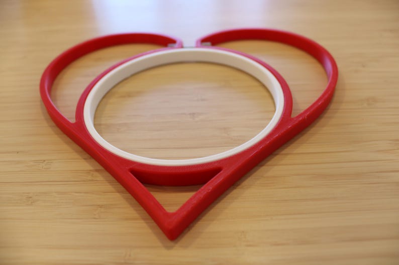May include: A red heart-shaped embroidery hoop frame with a white circular inner hoop. The red outer frame forms a distinct heart outline, while the white inner ring is positioned centrally to hold fabric securely within the heart-shaped structure.