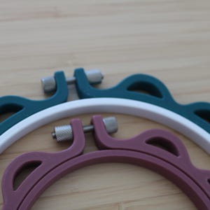 Op de afbeelding: Een set van drie in elkaar passende borduurringen met een donkergroene buitenring, een witte middelste ring en een mauve binnenring. Elke ring heeft een geschulpte buitenrand en een zilverkleurig metalen spanschroefmechanisme aan de bovenzijde.
