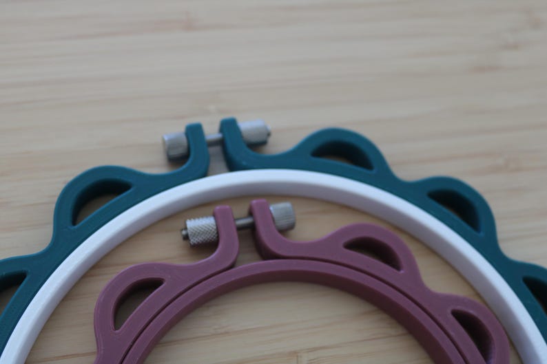 Op de afbeelding: Een set van drie in elkaar passende borduurringen met een donkergroene buitenring, een witte middelste ring en een mauve binnenring. Elke ring heeft een geschulpte buitenrand en een zilverkleurig metalen spanschroefmechanisme aan de bovenzijde.