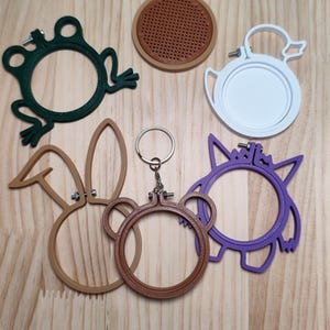 May include: A collection of six small embroidery hoops in various shapes and colors, including a dark green frog, a white duck, a purple creature, and three tan or brown animal-themed frames, one of which features a metal keychain ring.