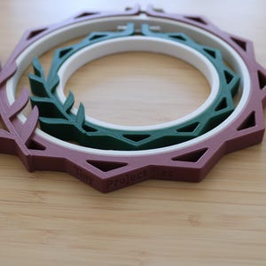 May include: A nested embroidery hoop set featuring a mauve outer frame with a leaf-like design, a white middle ring, and a dark green inner ring with a geometric pattern. The text Tiny Project lies is embossed on the mauve frame's edge.