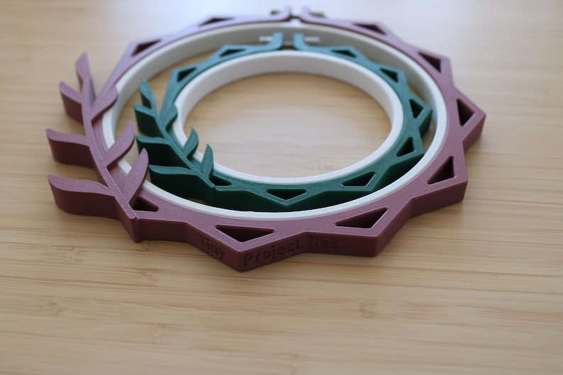 May include: A nested embroidery hoop set featuring a mauve outer frame with a leaf-like design, a white middle ring, and a dark green inner ring with a geometric pattern. The text Tiny Project lies is embossed on the mauve frame's edge.