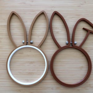 May include: Two circular embroidery hoops featuring attached rabbit ear shapes. The left hoop is light brown with a white inner ring, while the right hoop is dark brown with one ear folded down. Both hoops include small silver-toned metal tightening screws.