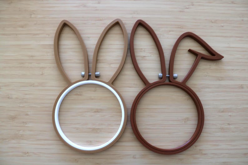 May include: Two circular embroidery hoops featuring attached rabbit ear shapes. The left hoop is light brown with a white inner ring, while the right hoop is dark brown with one ear folded down. Both hoops include small silver-toned metal tightening screws.