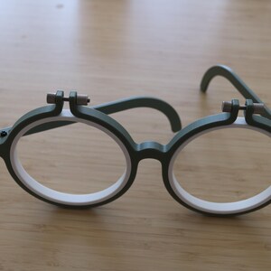 May include: A pair of round glasses featuring a dark green outer frame and a white inner rim. The top of each circular lens area includes a small, dark green adjustment screw mechanism with a metallic pin, resembling a miniature embroidery hoop.