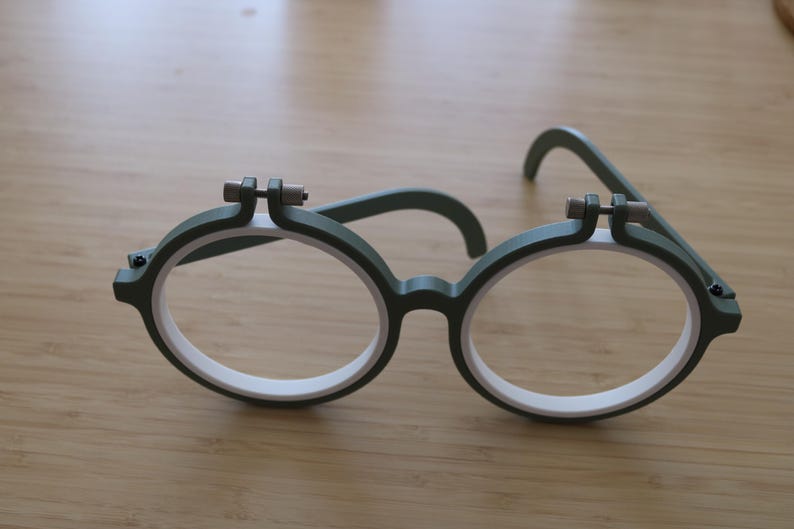 May include: A pair of round glasses featuring a dark green outer frame and a white inner rim. The top of each circular lens area includes a small, dark green adjustment screw mechanism with a metallic pin, resembling a miniature embroidery hoop.