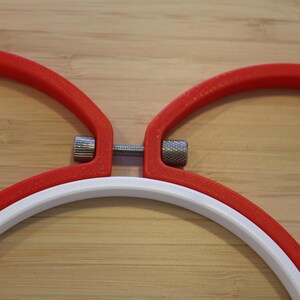 May include: Two red embroidery hoops and one white embroidery hoop arranged together. Each hoop features a metallic silver tension screw mechanism with a knurled texture, allowing for the adjustment of the hoop diameter to secure fabric.