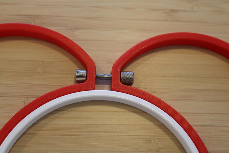 May include: Two red embroidery hoops and one white embroidery hoop arranged together. Each hoop features a metallic silver tension screw mechanism with a knurled texture, allowing for the adjustment of the hoop diameter to secure fabric.