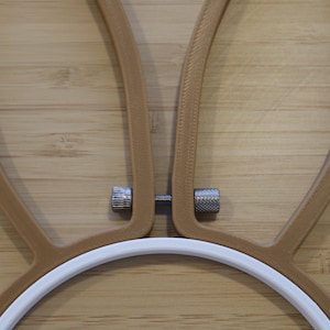 May include: An embroidery hoop featuring a white circular frame attached to two tan, ear-shaped extensions. A small, silver-colored metal tension screw with two knurled nuts connects the two tan extensions at the base of the ear shapes.