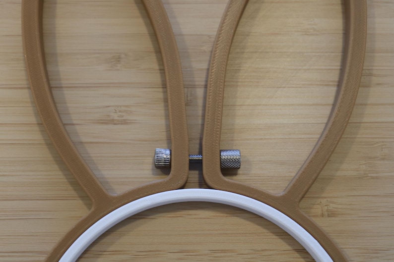 May include: An embroidery hoop featuring a white circular frame attached to two tan, ear-shaped extensions. A small, silver-colored metal tension screw with two knurled nuts connects the two tan extensions at the base of the ear shapes.