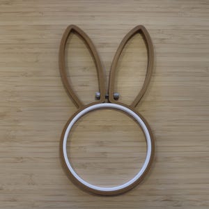 May include: A brown embroidery hoop shaped like a rabbit head with two long, upright ears and a circular base. The frame features a white inner ring and a small metal tension screw at the center between the ears to secure fabric in place.