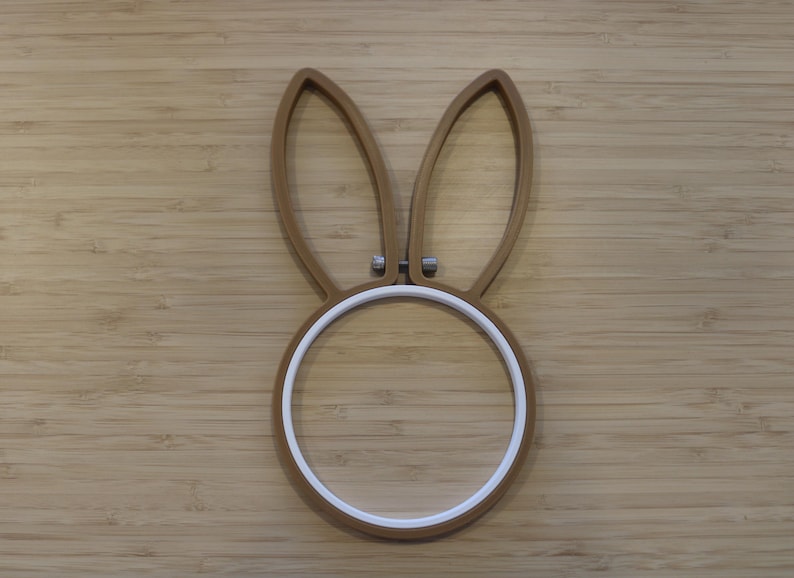 May include: A brown embroidery hoop shaped like a rabbit head with two long, upright ears and a circular base. The frame features a white inner ring and a small metal tension screw at the center between the ears to secure fabric in place.