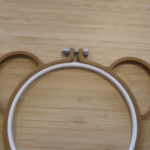 May include: A brown embroidery hoop shaped like a bear head with two circular ears on the sides. The hoop consists of an outer brown frame and an inner white ring, secured at the top by a silver-coloured metal tension screw for tightening fabric.