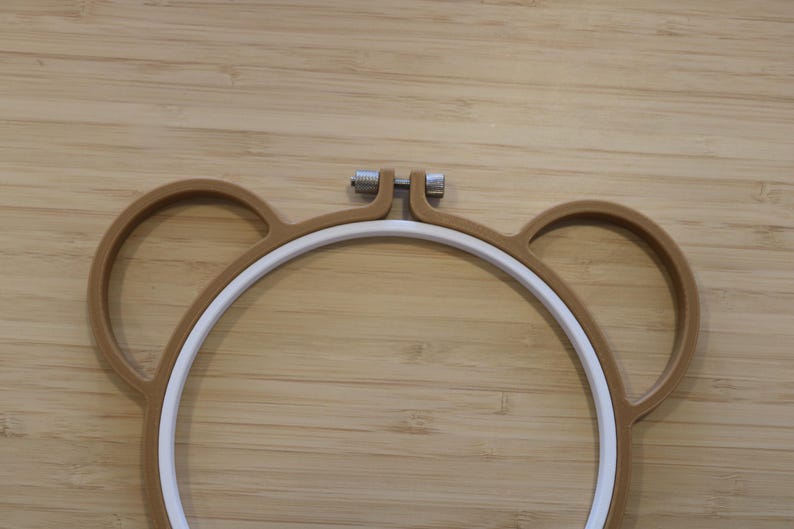 May include: A brown embroidery hoop shaped like a bear head with two circular ears on the sides. The hoop consists of an outer brown frame and an inner white ring, secured at the top by a silver-coloured metal tension screw for tightening fabric.