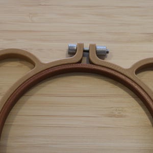 May include: A light brown embroidery hoop shaped like a bear head with two circular ears on the sides. The hoop features a central circular frame with a textured inner ring and a silver-coloured metal tightening screw at the top centre.