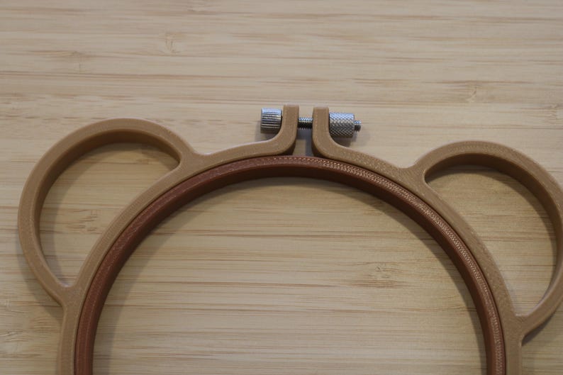 May include: A light brown embroidery hoop shaped like a bear head with two circular ears on the sides. The hoop features a central circular frame with a textured inner ring and a silver-coloured metal tightening screw at the top centre.
