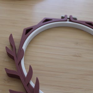 May include: A circular embroidery hoop featuring a white inner ring and a dark maroon outer frame. The outer frame is decorated with a series of triangular cutouts and a stylized leaf-like pattern along one side, secured by a metal screw at the top.