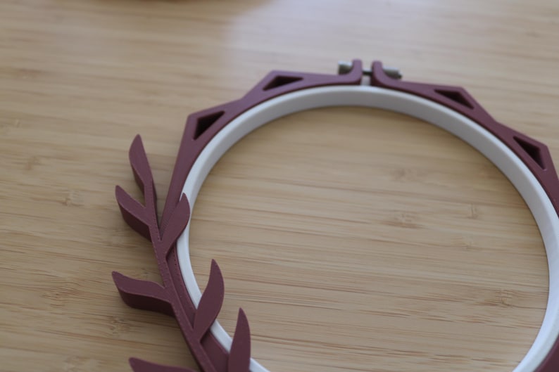 May include: A circular embroidery hoop featuring a white inner ring and a dark maroon outer frame. The outer frame is decorated with a series of triangular cutouts and a stylized leaf-like pattern along one side, secured by a metal screw at the top.