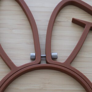 May include: A brown embroidery hoop shaped like rabbit ears. The frame consists of two connected pieces of brown plastic with a silver metal tension screw located between the two upright ear sections to secure fabric in place during needlework.