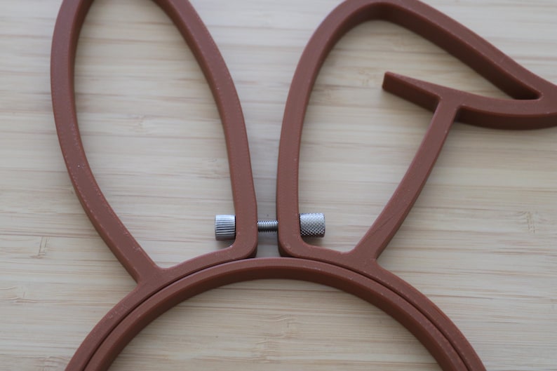 May include: A brown embroidery hoop shaped like rabbit ears. The frame consists of two connected pieces of brown plastic with a silver metal tension screw located between the two upright ear sections to secure fabric in place during needlework.
