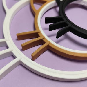 May include: Three circular frames shaped like cat faces, stacked on top of each other. The frames are white, tan, and black, each featuring two pointed ears at the top and three horizontal whiskers extending from the sides of the circular ring.