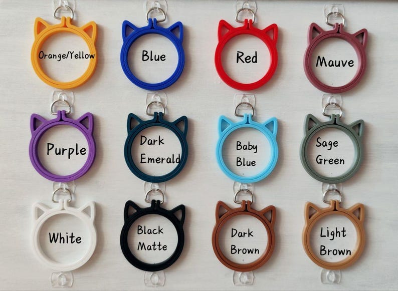 Op de afbeelding: Een collectie van twaalf ronde borduurringen in de vorm van kattenkoppen, elk voorzien van een metalen ophanglus. De kleuren zijn oranje/geel, blauw, rood, mauve, paars, donker smaragdgroen, babyblauw, saliegroen, wit, matzwart, donkerbruin en lichtbruin.