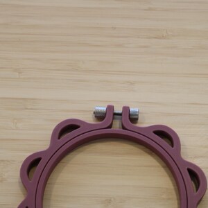 Op de afbeelding: Een cirkelvormige borduurring in een matte mauve kleur met een geschulpte buitenrand. De ring is voorzien van een zilverkleurig metalen spanschroefmechanisme aan de bovenzijde om het frame vast te zetten, gemaakt van stevig, glad materiaal.