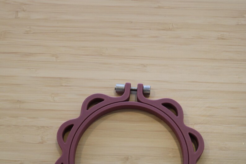 Op de afbeelding: Een cirkelvormige borduurring in een matte mauve kleur met een geschulpte buitenrand. De ring is voorzien van een zilverkleurig metalen spanschroefmechanisme aan de bovenzijde om het frame vast te zetten, gemaakt van stevig, glad materiaal.