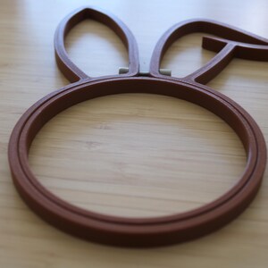 May include: A brown embroidery hoop featuring a circular frame with two attached rabbit ear shapes at the top. The hoop is constructed from a solid material with a smooth finish and includes a small metal tension adjustment mechanism at the base.