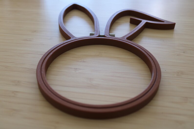 May include: A brown embroidery hoop featuring a circular frame with two attached rabbit ear shapes at the top. The hoop is constructed from a solid material with a smooth finish and includes a small metal tension adjustment mechanism at the base.