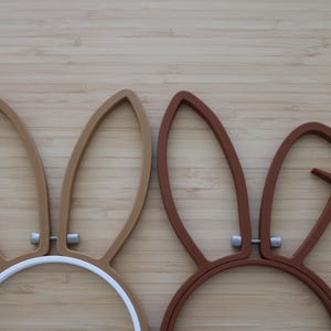 May include: Two embroidery hoops featuring attached rabbit ears. The left hoop is light brown with upright ears, while the right hoop is dark brown with one upright ear and one bent ear. Both hoops have silver-toned metal hardware at the base.