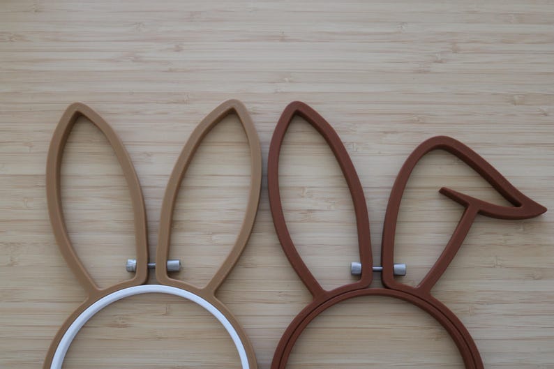 May include: Two embroidery hoops featuring attached rabbit ears. The left hoop is light brown with upright ears, while the right hoop is dark brown with one upright ear and one bent ear. Both hoops have silver-toned metal hardware at the base.