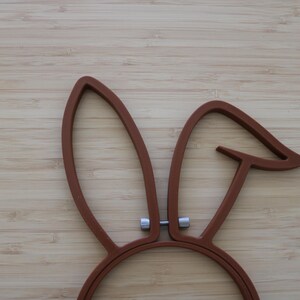May include: A brown embroidery hoop shaped like rabbit ears with a metallic silver tension screw at the base. The frame consists of two distinct ear-shaped sections, one standing upright and the other bent at an angle, connected by a central screw.