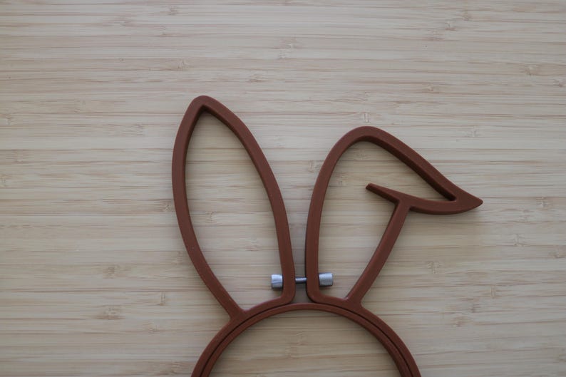 May include: A brown embroidery hoop shaped like rabbit ears with a metallic silver tension screw at the base. The frame consists of two distinct ear-shaped sections, one standing upright and the other bent at an angle, connected by a central screw.