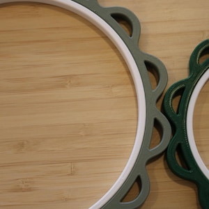 Op de afbeelding: Twee ronde borduurringen met witte binnenringen en geschulpte buitenframes. De grotere ring is saliegroen, terwijl de kleinere ring donker bosgroen is en aan de bovenkant is voorzien van een metalen schroef voor het spannen van de stof.