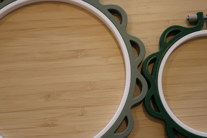 Op de afbeelding: Twee ronde borduurringen met witte binnenringen en geschulpte buitenframes. De grotere ring is saliegroen, terwijl de kleinere ring donker bosgroen is en aan de bovenkant is voorzien van een metalen schroef voor het spannen van de stof.