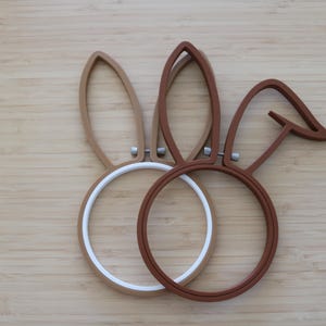 May include: Two circular embroidery hoops shaped like rabbit ears. One hoop is light tan with a white inner ring, and the other is dark brown. Each hoop features a small metal tightening screw at the base of the ears, creating a distinct silhouette.