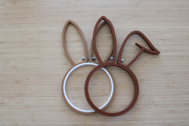 May include: Two circular embroidery hoops shaped like rabbit ears. One hoop is light tan with a white inner ring, and the other is dark brown. Each hoop features a small metal tightening screw at the base of the ears, creating a distinct silhouette.