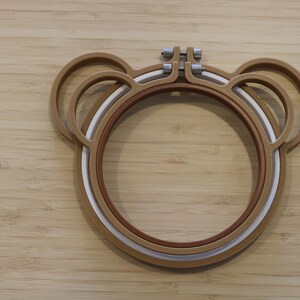 May include: A brown embroidery hoop shaped like a bear head with two circular ears on top. The hoop consists of an outer brown frame and an inner white frame, secured at the top by a metal screw mechanism for tightening the fabric during embroidery.