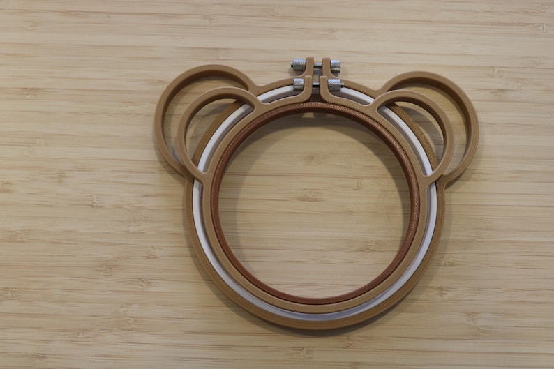 May include: A brown embroidery hoop shaped like a bear head with two circular ears on top. The hoop consists of an outer brown frame and an inner white frame, secured at the top by a metal screw mechanism for tightening the fabric during embroidery.