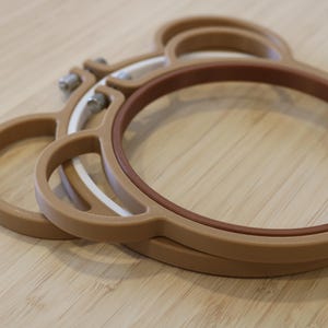 May include: A set of two tan embroidery hoops shaped like a bear head with rounded ears. The hoops feature a brown inner ring and a white inner ring, each secured with a metal tension screw at the top for adjusting the fabric tension during use.