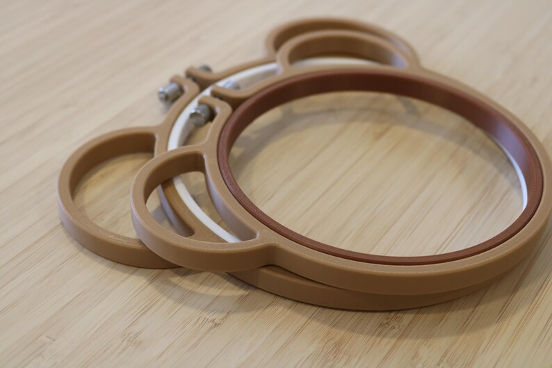 May include: A set of two tan embroidery hoops shaped like a bear head with rounded ears. The hoops feature a brown inner ring and a white inner ring, each secured with a metal tension screw at the top for adjusting the fabric tension during use.