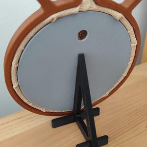 May include: A brown plastic embroidery hoop frame with a metal tightening screw at the top, holding a circular light gray backing panel. The frame is supported by a black plastic stand with a vertical support and a horizontal base.