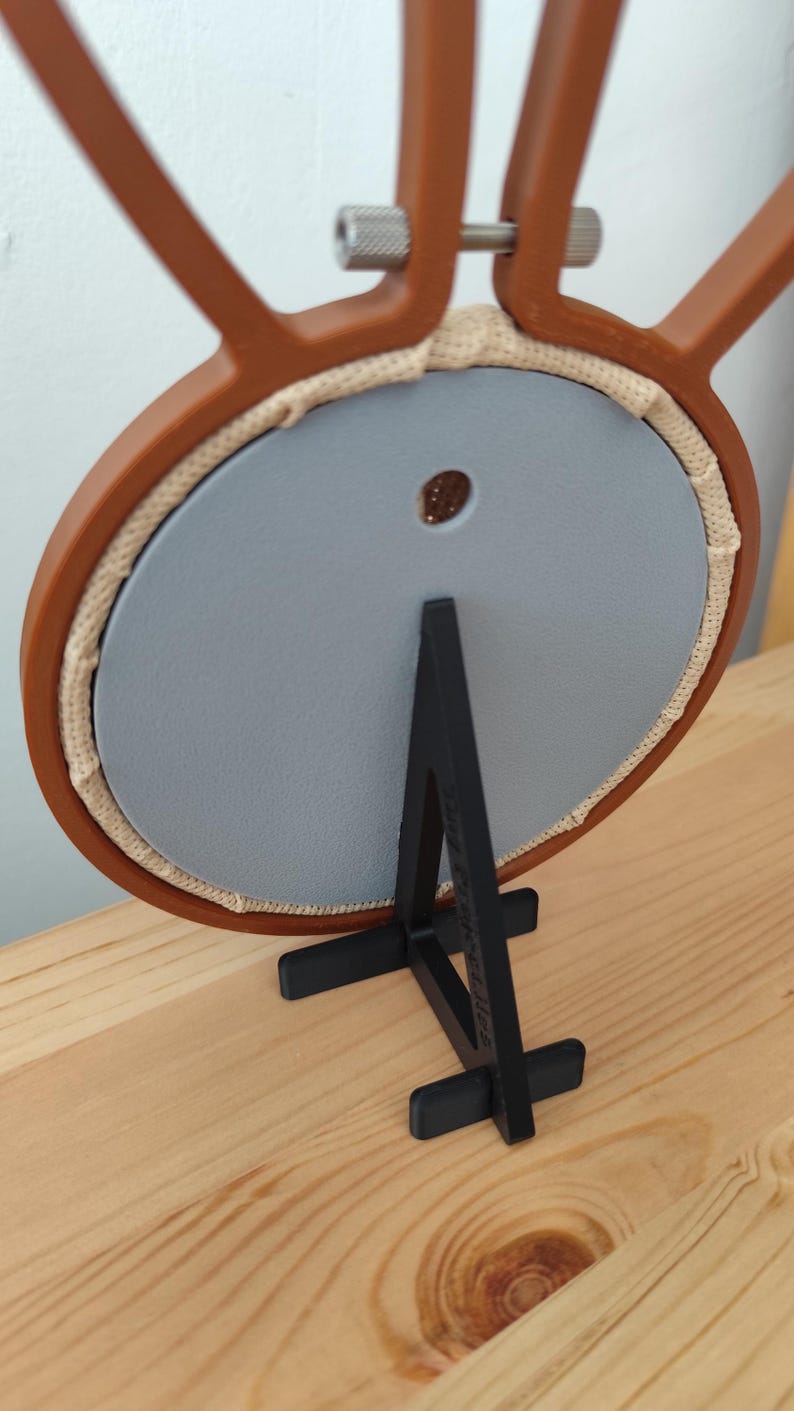 May include: A brown plastic embroidery hoop frame with a metal tightening screw at the top, holding a circular light gray backing panel. The frame is supported by a black plastic stand with a vertical support and a horizontal base.