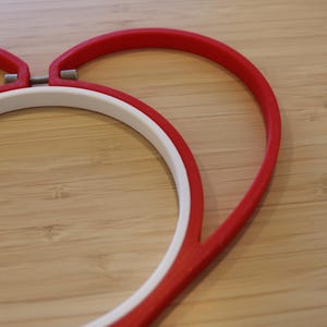May include: A heart-shaped embroidery hoop featuring a vibrant red outer frame and a circular white inner ring. The two components are connected by a small metal tension screw at the top center, designed to hold fabric securely in place for stitching.