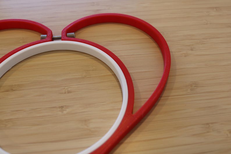 May include: A heart-shaped embroidery hoop featuring a vibrant red outer frame and a circular white inner ring. The two components are connected by a small metal tension screw at the top center, designed to hold fabric securely in place for stitching.