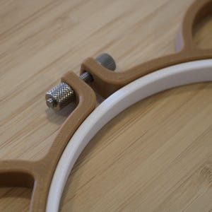 May include: An embroidery hoop featuring a white inner ring and a tan outer frame. The outer frame is secured with a metallic silver-coloured knurled tension screw mechanism that allows for tightening and loosening the hoop during use.