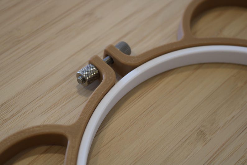 May include: An embroidery hoop featuring a white inner ring and a tan outer frame. The outer frame is secured with a metallic silver-coloured knurled tension screw mechanism that allows for tightening and loosening the hoop during use.