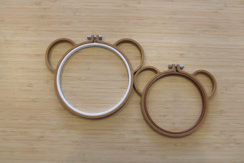 May include: Two circular embroidery hoops shaped like bear heads with rounded ears. The larger hoop features a white inner ring, while the smaller hoop has a brown inner ring. Both hoops include a metal tightening screw at the top centre.