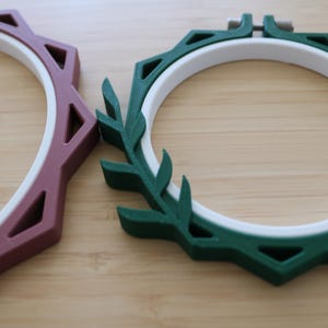 May include: Two circular embroidery hoops featuring white inner rings and colored outer frames. One frame is dark green with a leaf-like design on one side, and the other is a muted mauve with a geometric, angular, and faceted outer edge.